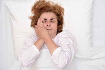 Mature woman with toothache lying in bed and holding her cheek. Top view.