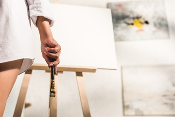 cropped view of girl holding paintbrush near easel