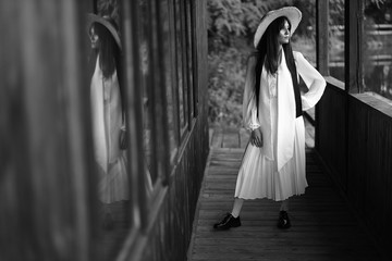 Fototapeta na wymiar Monochrome portrait of young beautiful woman in white dress and big straw hat, standing near wooden building