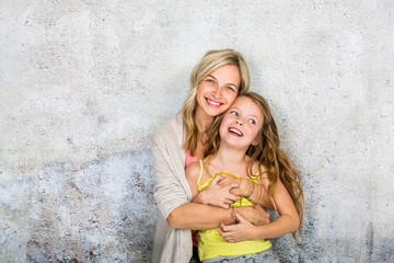 pretty, blonde young mother poses and cuddles with her daughter in front of a concrete wall and...