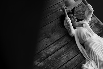 Black and white dreamy portrait of woman in vintage clothes and straw hat, lying on pier near pond.