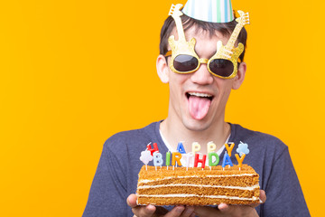 Crazy cheerful young asian guy with glasses holding a burning candle in his hands and a congratulatory homemade cake on a yellow background. Birthday and anniversary celebration concept.