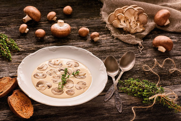 Plate with mushroom cream soup, pieces of toasted bread, melchior spoons, bundle of fresh thyme, brown royal champignons and oyster mushrooms on wooden table. Low fat food concept. Warm colors