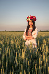 Beautiful indian young woman in traditional Ukrainian embroidery clothes and floral handmade wreath stand in field, look in distance dreaming or thinking, pretty ethnic woman posing in nature 