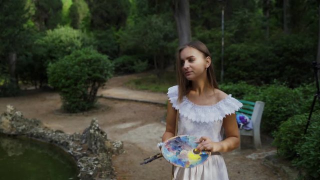 Girl artist prepares paper palette and paint brushes for landscape painting in the park with a small pond 4k
