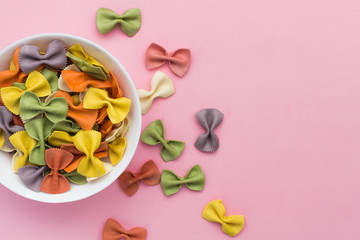Italian raw multicolored farfalle pasta close-up in a bowl on pink background. Colorful pasta in bow tie shape. Italian cuisine concept. Copy space.