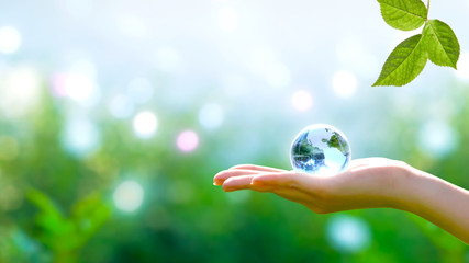 Earth crystal blue glass globe in human hand and fresh green leaf on blurred sky and trees background. Saving environment and clean green planet concept. Card for World Earth Day concept.