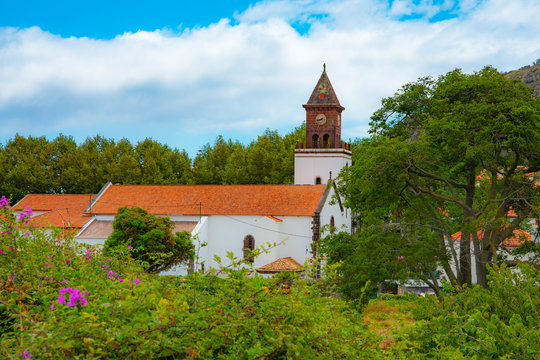 Urban landscape. There is a small Catholic Church under an orange tiled roof. There is a chapel next to the Church. The background images are green vegetation, blue sky and white clouds.