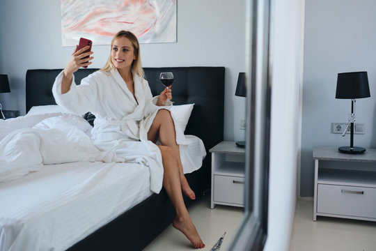 Young pretty smiling blond woman in white bathrobe holding glass of red wine in hand happily taking photo on cellphone sitting on bed in modern hotel