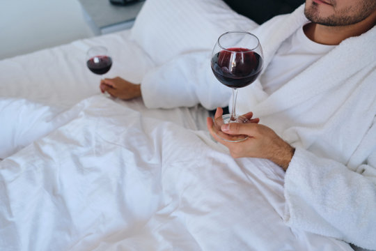 Close up photo of young attractive man in white bathrobe lying in bed with glasses of red wine in hands in modern hotel