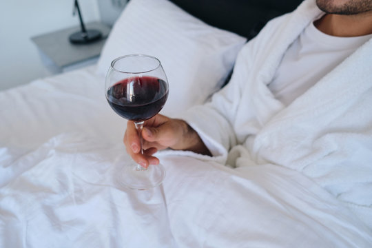 Close up photo of young attractive man in white bathrobe lying in bed with glass of red wine in hand in hotel