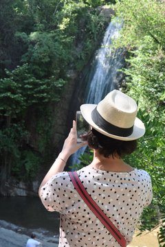 A young woman in a white hat, a pink T-shirt and with a bag over her shoulder takes pictures of a waterfall in the mountains to the phone