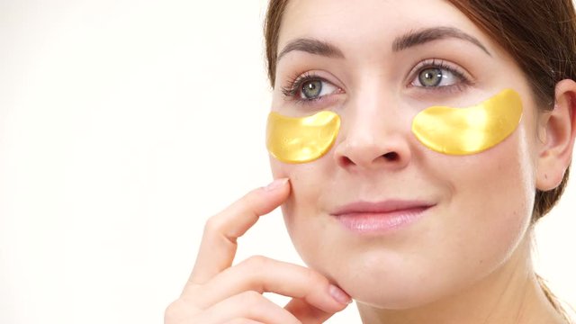 Young woman applying golden collagen patches under eyes, against white. Mask removing wrinkles and dark circles. Girl taking care of delicate skin around eye. Beauty treatment