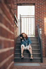 Beautiful red-haired girl posing with a camera on the street near a brick building on a summer evening