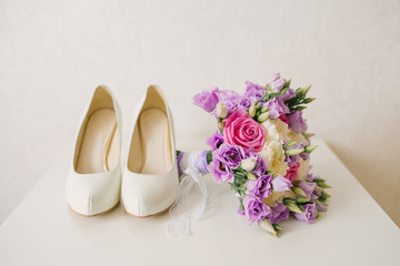 The bride's shoes and pink lilac wedding bouquet lies next to the white background, the bride's accessories
