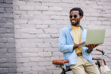 smiling african american man looking away while standing by brick wall near bicycle and using laptop