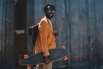 stylish african american businessman holding longboard and smiling at camera