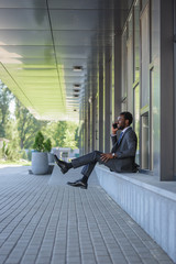 cheerful african american businessman talking on smartphone while sitting near office building