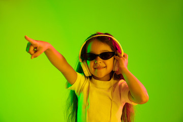 Beautiful female half-length portrait isolated on green backgroud in neon light. Young emotional teen girl. Human emotions, facial expression concept. Dancing in sunglasses and pointing up.