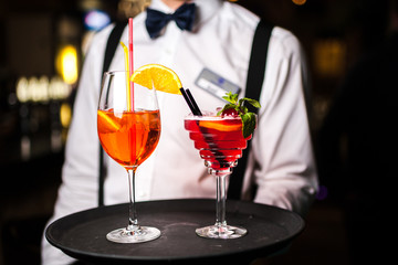 The waiter serves two cocktails on a tray. Close-up orange and red cocktails with orange and mint.