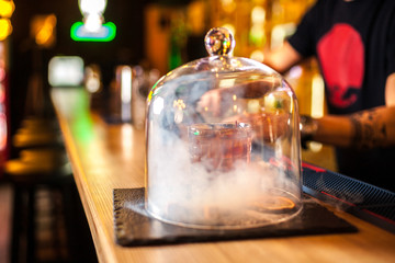 Close-up cocktail with cinnamon and raisins on the bar, blurred background. Cocktail under the hood with smoke effect.