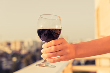 Woman hand with glass of red wine against the city background at summer evening