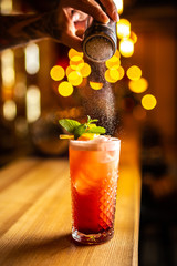 Bartender finishes preparation of orange cocktail in crystal glass by adding a bitter of powdered sugar. Close-up of expert bartender making cocktail in bar.