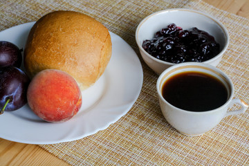 Homemade breakfast, cup of coffee, bun and fruit on a plate, peach and plums, jam in a bowl