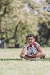 cute, bored schoolgirl holding book while sitting on lawn in park