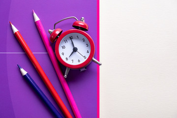 Colorful pencils lying on a purple notebook and red alarm clock. Back to school concept. Time management. Top view