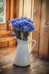 Flowers of a cornflower in a white iron pitcher. The pitcher stands on a wooden shelf. A bouquet of flowers in a jug. Blue flowers in a vase.