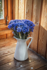 Flowers of a cornflower in a white iron pitcher. The pitcher stands on a wooden shelf. A bouquet of flowers in a jug. Blue flowers in a vase.