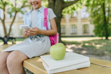 Fototapeta premium cropped view of smiling schoolgirl using smartphone while sitting on bench near books and apple