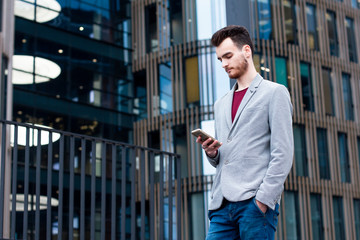 Businessman with mobile phone outside office. Young urban professional man using smart phone. Guy wearing gray jacket, with a beard, holding mobile smartphone, texting sms message. Workplace, outdoor.