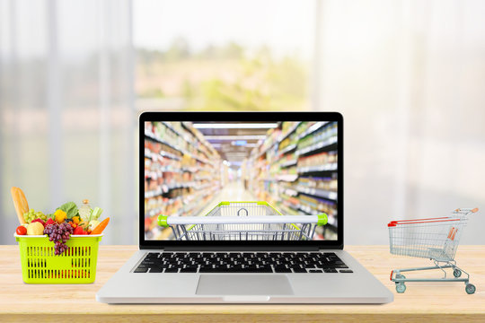 laptop computer and shopping cart on wood table with window curtain abstract blur background grocery online concept