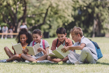Four multicultural schoolkids sitting on lawn and reading books