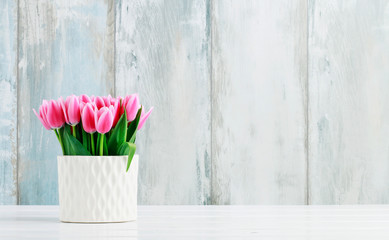Pink tulips in white ceramic vase, wooden wall background.