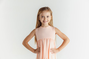 Stylish little smiling girl posing in dress isolated on white studio background. Caucasian blonde female model. Human emotions, facial expression, childhood. Smiling, holding hands on a belt.