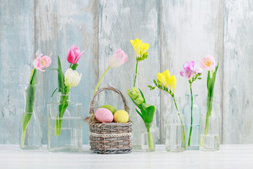 Colorful flowers in glass bottles and Easter eggs in wicker basket on the table