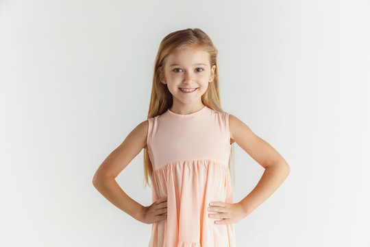 Stylish little smiling girl posing in dress isolated on white studio background. Caucasian blonde female model. Human emotions, facial expression, childhood. Smiling, holding hands on a belt.