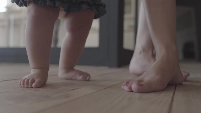 Close-up feet of a young woman and her baby standing on the floor at home. Concept of a happy family, one child, love. Slow motion