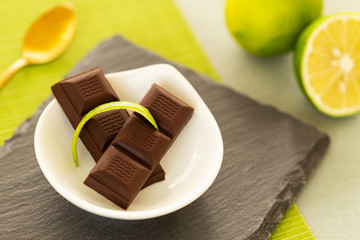 Chocolate pieces with lime peel in a small bowl on a slate, with a lime and a golden spoon over a green table mat and a light green tablecloth background. Closeup.