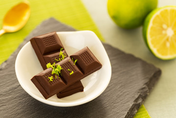 Chocolate pieces with shredded lime peel in a small bowl on a slate, with a lime and a golden spoon over a green table mat and a light green tablecloth background. Closeup.