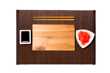 Empty rectangular brown wooden plate with chopsticks for sushi, ginger and soy sauce on dark bamboo mat background. Top view with copy space for you design