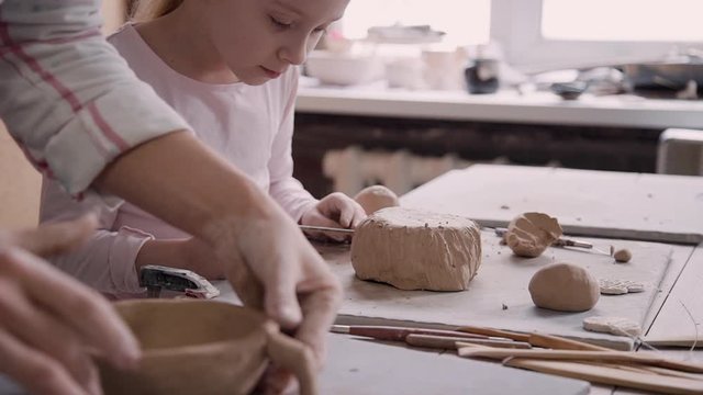 Adult ceramist person standing near little girl child in cozy class room or work place studio with soft light inside. Two people working with raw clay and making new beautiful objects
