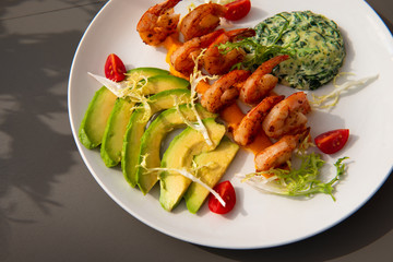 Healthy shrimp dish. Delicious grilled shrimps with avocado and salad on a white plate. Food decorated with cherry tomatoes. Sunlight.