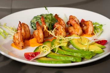 Healthy shrimp dish. Delicious grilled shrimps with avocado and salad on a white plate. Food decorated with cherry tomatoes. Sunlight.