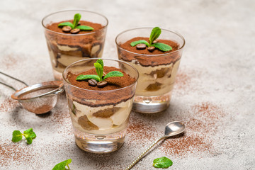 Classic tiramisu dessert in a glass cup and strainer with cocoa powder on concrete background
