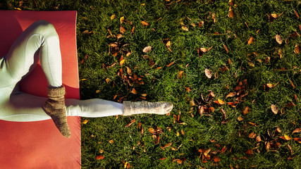 Yoga girl in autumn park with yoga mat on green carpet with fallen yellow leaves. Funny autumn legs wearing woolen socks and doing yoga in cold autumn park in the morning