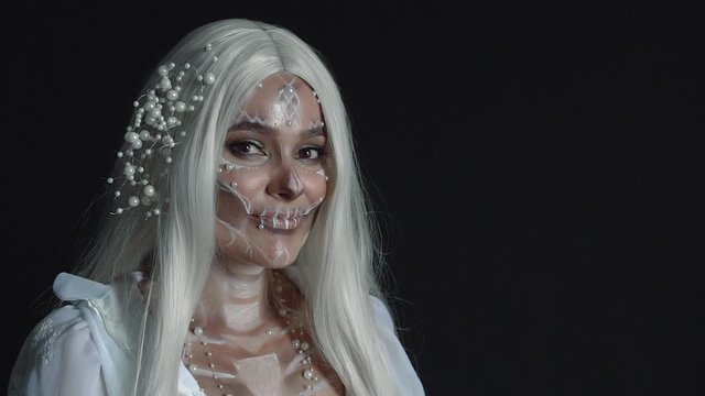 charming sweet girl with sweet skull painted on face, happy cheerful death goddess with white hair stands in black room and playfully winks, Mexican celebration for dead, welcomes souls of departed